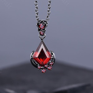 May include: A pendant necklace featuring a dark metal chain and a striking red gemstone. The pendant has a teardrop shape with a faceted red stone, accented by smaller red gems. The necklace is a piece of jewelry.