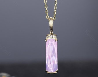 May include: A gold-colored necklace with a rectangular, light pink pendant. The pendant is faceted and has a small, diamond-studded cap. The chain is delicate and hangs against a dark background. The necklace is a piece of jewelry.
