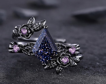 May include: A black ring set featuring a geometric blue sandstone center stone. The ring is adorned with delicate floral and leaf accents, and small pink gemstones. The ring is set against a dark, textured background.