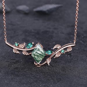 May include: A rose gold necklace with a central green gemstone, square-cut, and smaller green stones, with leaf details. The necklace has a fine chain and a nature-inspired design, ideal for adding a touch of elegance to any outfit.