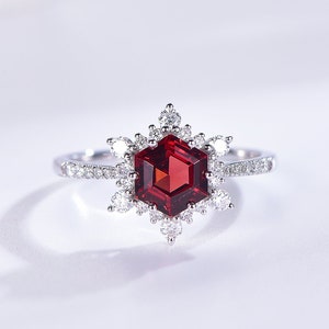 May include: A silver ring featuring a red gemstone surrounded by smaller white gemstones. The ring has a snowflake-like design.