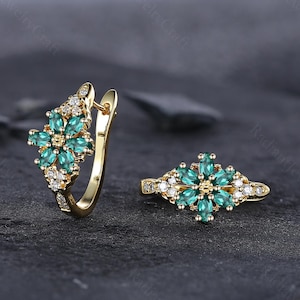 Op de afbeelding: Een paar gouden oorbellen en een bijpassende ring. De sieraden hebben een bloemmotief met smaragdgroene edelstenen en kleine witte diamanten. De oorbellen zijn van het creoolse type en de ring heeft een vergelijkbare bloemenopstelling.