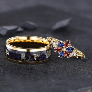 Puede incluir: Dos anillos de oro. Uno tiene una banda azul oscuro con detalles plateados y rojos. El otro anillo tiene una banda dorada con un diseño floral con piedras preciosas rojas, azules y blancas. Los anillos están sobre un fondo oscuro.
