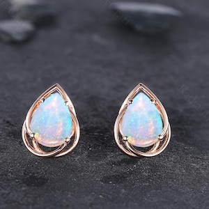 May include: A pair of rose gold teardrop stud earrings with iridescent opal stones. The earrings have a delicate, openwork design around the opal, set against a dark gray background.