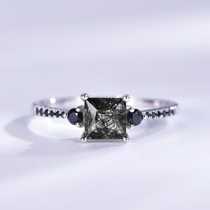 May include: A silver ring featuring a square black gemstone, flanked by two smaller black gemstones. The band is adorned with a row of tiny black gemstones.