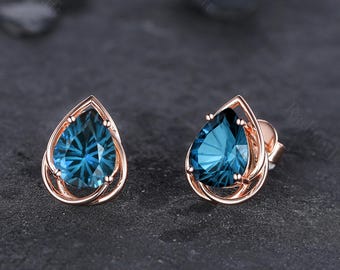 Könnte beinhalten: Ohrstecker aus Roségold mit tropfenförmigen, teal-farbenen Edelsteinen. Die Edelsteine sind in einem filigranen, offenen Rahmen gefasst. Die Ohrringe werden auf einer dunklen, strukturierten Oberfläche präsentiert.