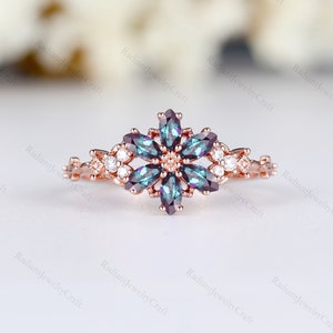 Könnte beinhalten: Ein Ring aus Roségold mit einem blütenförmigen Design, das einen zentralen Stein zeigt, der von kleineren Steinen umgeben ist. Der zentrale Stein ist ein dunkelblaugrüner Edelstein mit einem Regenbogeneffekt. Die kleineren Steine sind klar und rund.