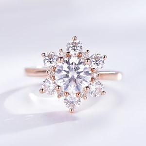 Könnte beinhalten: Ein roségoldener Ring mit einem großen, runden, klaren Edelstein, der von kleineren, klaren Edelsteinen umgeben ist und ein Stern- oder Schneeflockenmuster bildet. Der Ring hat ein schlichtes, dünnes roségoldenes Band.