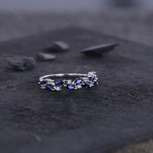 Peut inclure: Une bague en argent avec un motif floral. La bague pr&eacute;sente des saphirs bleus taille marquise et de petits diamants blancs ronds. Les pierres pr&eacute;cieuses sont serties dans un motif d&eacute;licat en forme de feuille. La bague est pr&eacute;sent&eacute;e sur une surface sombre et textur&eacute;e.