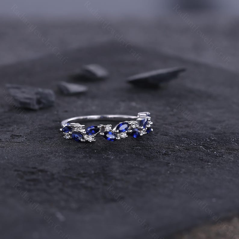 Peut inclure: Une bague en argent avec un motif floral. La bague pr&eacute;sente des saphirs bleus taille marquise et de petits diamants blancs ronds. Les pierres pr&eacute;cieuses sont serties dans un motif d&eacute;licat en forme de feuille. La bague est pr&eacute;sent&eacute;e sur une surface sombre et textur&eacute;e.