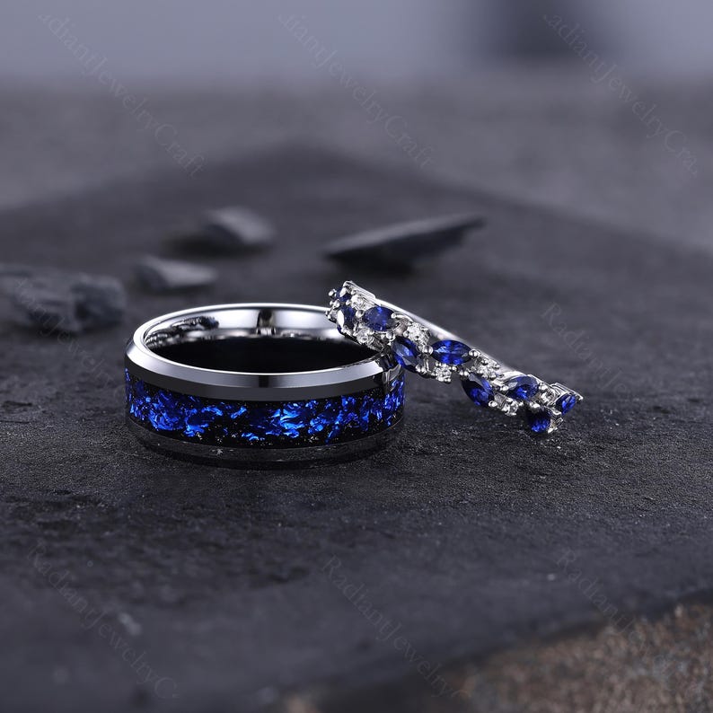 Peut inclure: Deux bagues, l'une avec une large bande orn&eacute;e d'un incrustation mouchet&eacute;e bleue et noire, et l'autre avec une bande plus fine orn&eacute;e de pierres pr&eacute;cieuses bleues et de petites pierres blanches. Les bagues sont pos&eacute;es sur un fond sombre et textur&eacute;.