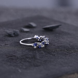 Peut inclure: Une bague en argent orn&eacute;e d'une s&eacute;rie de pierres bleues en forme de marquise et de petites pierres blanches. La bague est pos&eacute;e sur un fond sombre et textur&eacute;, mettant en valeur l'&eacute;clat des pierres pr&eacute;cieuses. Le design de la bague est d&eacute;licat et &eacute;l&eacute;gant.