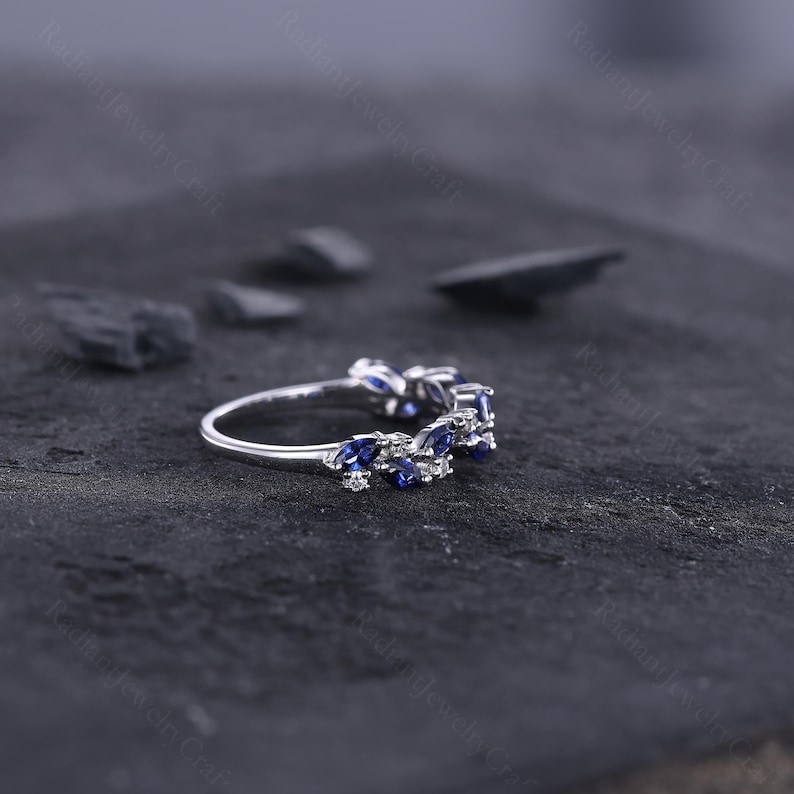 Peut inclure: Une bague en argent orn&eacute;e d'une s&eacute;rie de pierres bleues en forme de marquise et de petites pierres blanches. La bague est pos&eacute;e sur un fond sombre et textur&eacute;, mettant en valeur l'&eacute;clat des pierres pr&eacute;cieuses. Le design de la bague est d&eacute;licat et &eacute;l&eacute;gant.
