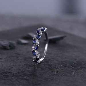 Peut inclure: Une bague en argent orn&eacute;e d'une rang&eacute;e altern&eacute;e de pierres pr&eacute;cieuses bleu fonc&eacute; de taille marquise et de petites pierres claires. La bague est pr&eacute;sent&eacute;e sur un fond sombre et textur&eacute;, mettant en valeur l'&eacute;clat des gemmes. Le design de la bague est &eacute;l&eacute;gant.