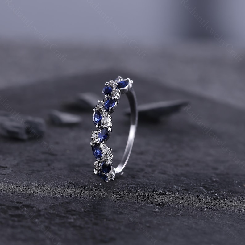 Peut inclure: Une bague en argent orn&eacute;e d'une rang&eacute;e altern&eacute;e de pierres pr&eacute;cieuses bleu fonc&eacute; de taille marquise et de petites pierres claires. La bague est pr&eacute;sent&eacute;e sur un fond sombre et textur&eacute;, mettant en valeur l'&eacute;clat des gemmes. Le design de la bague est &eacute;l&eacute;gant.