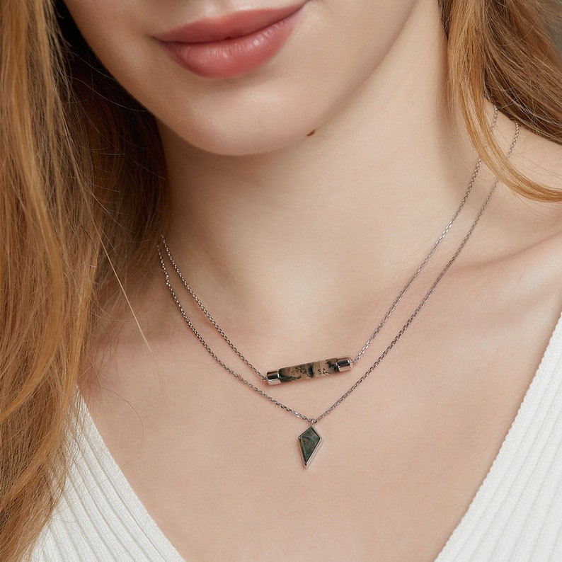 May include: A silver chain necklace with two pendants. The top pendant is a long, thin bar with a green and brown stone. The bottom pendant is a small, diamond-shaped pendant with a green and brown stone.