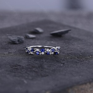 Peut inclure: Une bague en argent orn&eacute;e d'une rang&eacute;e de pierres pr&eacute;cieuses bleues en forme de marquise et de petites pierres blanches. La bague est pos&eacute;e sur un fond sombre et textur&eacute;, avec quelques pierres grises fonc&eacute;es &eacute;parpill&eacute;es autour.