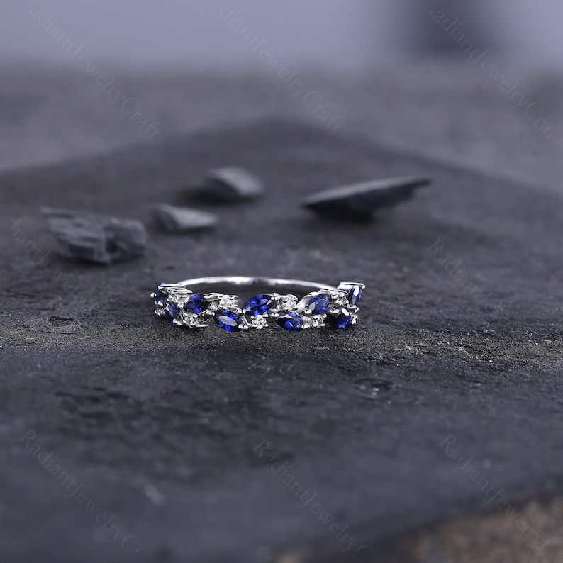 Peut inclure: Une bague en argent orn&eacute;e d'une rang&eacute;e de pierres pr&eacute;cieuses bleues en forme de marquise et de petites pierres blanches. La bague est pos&eacute;e sur un fond sombre et textur&eacute;, avec quelques pierres grises fonc&eacute;es &eacute;parpill&eacute;es autour.