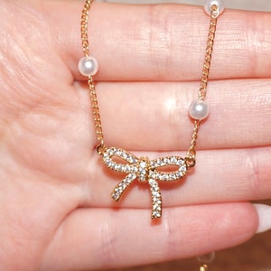 May include: A gold chain necklace with a sparkling bow pendant and white pearl accents.