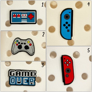 May include: Five different video game themed shoe charms. The charms are made of rubber and feature a retro video game console, a gray video game controller, a blue Nintendo Switch controller, a red Nintendo Switch controller, and a blue and red pixelated "GAME OVER" sign.