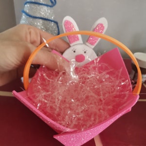 Può includere: Un cestino di Pasqua rosa e arancione con un viso di coniglio bianco e glitter rosa. Il cestino è pieno di erba di plastica trasparente.