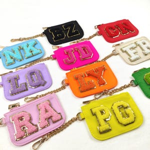 May include: A selection of small coin purses in various colours, each with a gold chain strap. Each purse displays a different letter combination in a textured, embroidered style. Colours include black, pink, orange, and yellow.