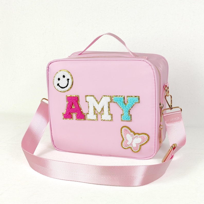 May include: Pink lunch bag with a shoulder strap and top handle. The bag features the name "AMY" in colorful, glittery letters, a smiley face patch, and a butterfly patch. The bag has gold-colored hardware.