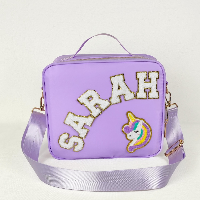 May include: A lavender crossbody bag with the name "SARAH" in white and gold letters. A unicorn patch is on the front. The bag has a top handle and a lavender adjustable strap with gold-colored hardware.