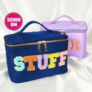 May include: Two cosmetic bags. One is navy blue with the word "STUFF" in colorful, textured letters. The other is lavender with the word "HER". Both bags have gold zippers and handles. A pink circle with the words "SEWN ON" is also visible.