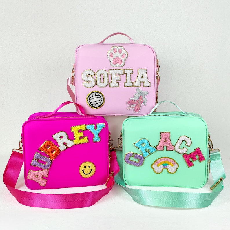 May include: Three colorful square crossbody bags. One is pink with the name "SOFIA", a paw print, and a ballet shoe. Another is hot pink with the name "AUBREY" and a smiley face. The third is mint green with the name "GRACE" and a rainbow.