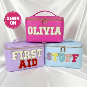 May include: Three colorful cosmetic bags with gold zippers and handles. The pink bag has the name "OLIVIA" in white letters. The purple bag says "FIRST AID" in white and red letters. The blue bag says "STUFF" in colorful letters.