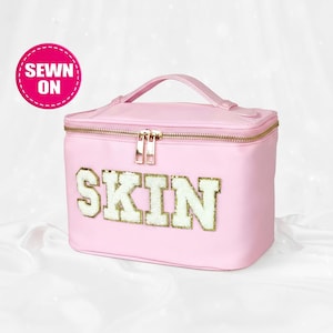 May include: A pink cosmetic bag with a gold zipper and handle. The word "SKIN" is embroidered on the front in white and gold letters. A pink circle with the words "SEWN ON" is in the upper left corner.