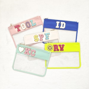 May include: Five clear plastic zipper pouches with colorful fabric flaps. The flaps have different letters and designs, including "TOOL", "ID", "SPF", "KC" with a heart, and "RV" with a smiley face.