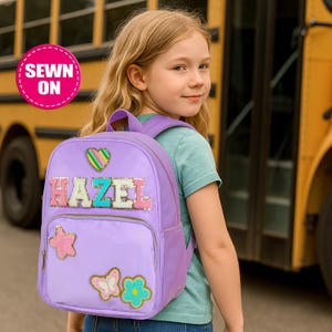 Peut inclure: Un sac à dos lavande avec le nom "HAZEL" en lettres à paillettes. Le sac à dos comporte des ornements en forme de cœur, d'étoile, de papillon et de fleur. Les mots "SEWN ON" sont dans un cercle rose en haut à gauche. Le sac à dos est sur le dos d'une personne.