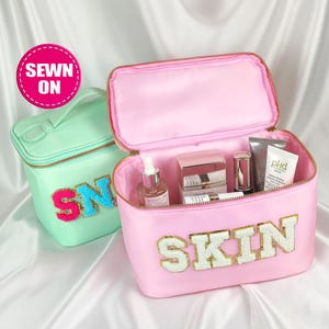 Puede incluir: Dos neceseres, uno verde menta y otro rosa, con cremalleras doradas. El neceser rosa está abierto, revelando productos para el cuidado de la piel. El neceser rosa tiene la palabra "SKIN" en letras doradas y blancas. El neceser verde menta tiene las letras "SN" en rosa y azul.