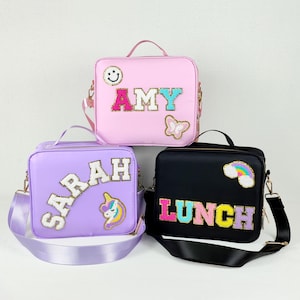 May include: Three lunch bags in pink, purple, and black. The pink bag has the name "AMY" in colorful letters, a smiley face, and a butterfly. The purple bag has the name "SARAH" and a unicorn. The black bag says "LUNCH" with a rainbow.