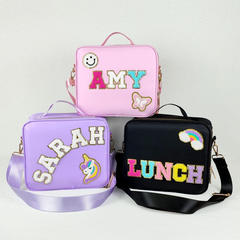 May include: Three lunch bags in pink, purple, and black. The pink bag has the name "AMY" in colorful letters, a smiley face, and a butterfly. The purple bag has the name "SARAH" and a unicorn. The black bag says "LUNCH" with a rainbow.