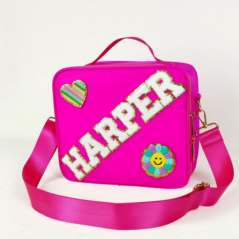May include: Bright pink lunch bag with a matching shoulder strap and top handle. The bag features the name "HARPER" in white, embroidered letters, along with a heart and flower patch. The bag has gold-colored hardware.