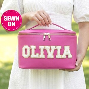 May include: A bright pink rectangular cosmetic bag with the name "OLIVIA" in white and gold letters. The bag has a gold zipper and a handle. A pink circle with the words "SEWN ON" is in the upper left corner.