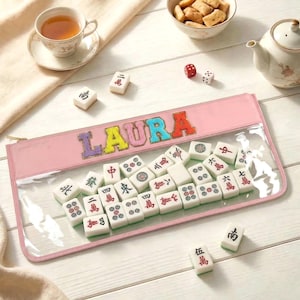 May include: A pink zippered pouch with a clear front panel containing white Mahjong tiles. The name LAURA is appliqued in colorful fabric letters on the pink section. The pouch is surrounded by loose Mahjong tiles, a teacup, a teapot, and a bowl of snacks.