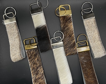 May include: Seven keychains made of brown and white cowhide with silver and gold metal rings. The keychains are all different lengths and have different patterns.