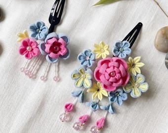 May include: Two floral hair clips with black bases. Each clip features a cluster of fabric flowers in pink, blue, and yellow, with pearl accents. The clips have dangling beaded strands and are set against a white background.