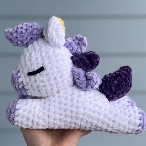 May include: A white and purple crocheted unicorn plush toy with a yellow horn. The unicorn is lying on its side with its eyes closed.