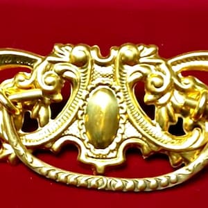 May include: A gold-toned ornate drawer pull with a decorative design featuring a central oval and intricate scrollwork.