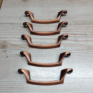 May include: Five bronze-colored cabinet pulls with a curved handle design and decorative scroll ends. The pulls are arranged on a light wood-grain surface, showcasing their antique style and potential for furniture restoration projects.
