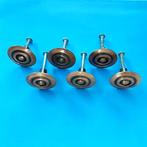 May include: Six antique copper-toned cabinet knobs with a concentric circle design. Each knob has a central screw post and a circular face with a series of raised rings. The knobs are set against a bright blue background.