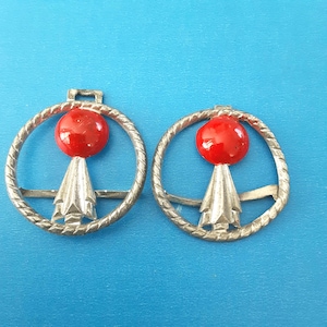 May include: Two vintage silver-toned metal brooches with a red sphere and a stylized arrow design. The brooches have a circular, rope-like border and a small loop at the top for attachment. The background is a solid blue.