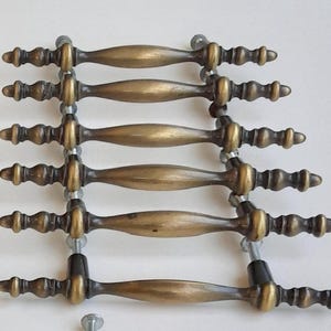 May include: A set of antique brass cabinet handles. Each handle features a central, oval-shaped grip with decorative detailing at each end. The handles are mounted on metal rods with screws, ready for installation. The handles have a warm, aged patina.