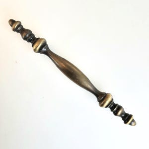 May include: A bronze-toned cabinet pull with a decorative design. The handle has a central grip and ornate ends with a series of rounded shapes. The hardware is set against a white background.