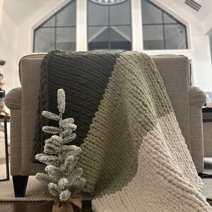 May include: A chunky knit blanket in olive green, sage green, and cream colors draped over a gray armchair. A small, frosted Christmas tree sits in front of the blanket. The blanket is a cozy home decor item.